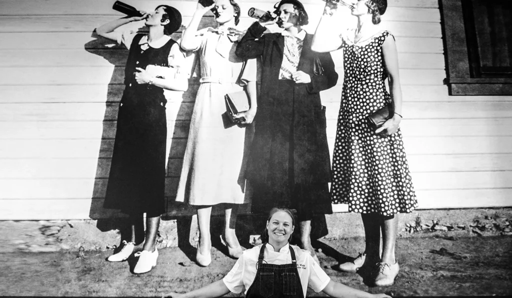Chef Sarah Wade standing in front of a prohibition era mural of women drinking alcohol